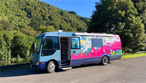 Image shows the mobile library bus of Upper Hutt Libraries parked on the road. Native forest on Upper Hutt's surrounding hills is in the background.