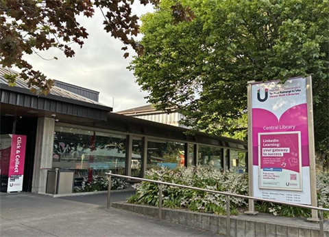 Image shows the front view of the Central Library in Upper Hutt. The building is surrounded by greenery and trees. Pictured on the left is the pink Click and Collect locker.